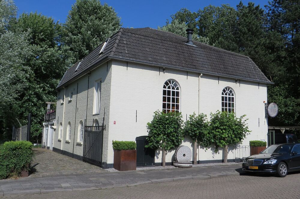 Wit geschilderd pand met hoge rondboogramen en een pannendak aan de Molenstraat in Boxtel, met leibomen langs de gevel, een oude molensteen tegen de muur en een diepblauwe lucht erboven